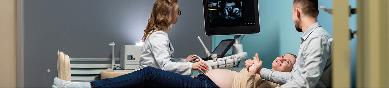 De foto toont een zwangere vrouw liggend op een stoel. Links van haar staat een vrouw in een witte jas die een echo maakt van de baby. Boven de zwangere vrouw hangt een monitor. Rechts van de zwangere vrouw staat een man die haar hand vasthoudt.