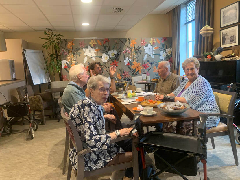 De foto toont 4 oudere mensen aan tafel en 1 jongere man op de achtergrond. Op de voorgrond staat een rollator.