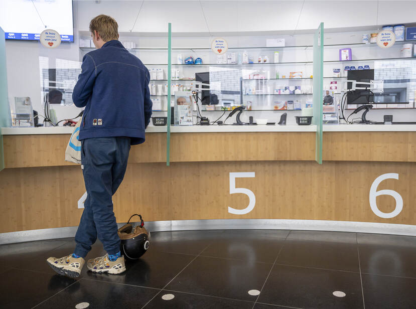 De foto toont een jongeman die aan de balie van een apotheek staat. We zien hem op de rug. Hij heeft blond haar, draagt blauwe kleding en witte gympen. Aan zijn voeten ligt een scooterhelm. De lichthouten balie heeft 3 loketten met de nummers 4, 5, en 6. De jongen staat bij 4. De andere loketten zijn leeg.