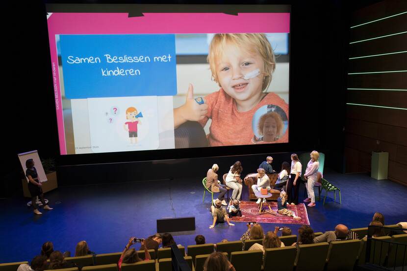 Op de foto zien we het podium van de conferentie samen beslissen met kinderen. De foto is gemaakt vanuit de zaal. Op het podium zien we een aantal volwassen en kinderen die omkijken naar een presentatie die wordt getoond op een groot scherm.